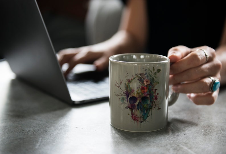 Puede incluir: Una taza de cer&aacute;mica gris con un dise&ntilde;o de calavera acuarela rodeada de flores coloridas. La taza est&aacute; siendo sostenida por una mano que lleva anillos.