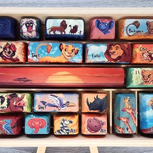May include: A wooden box containing custom keycaps with Lion King illustrations. The keycaps depict characters such as Simba, Timon, Pumbaa, and Scar, alongside keyboard function labels. The colour scheme includes shades of orange, blue, and brown.
