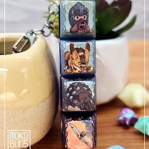 May include: A keychain with four illustrated cubes stacked together. Each cube shows a different fantasy creature or scene, with a blue and white colour scheme. The keychain has a silver clasp and a small logo that says "Moko Buns".