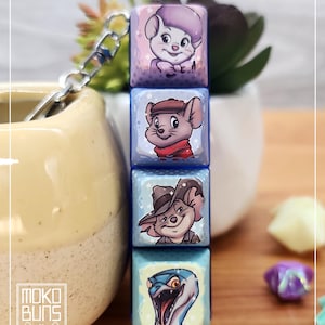 May include: A stacked keychain with four square charms, each displaying a cartoon mouse or snake illustration. The charms are in blue, purple, and brown. The keychain is attached to a silver chain. The background is blurred.