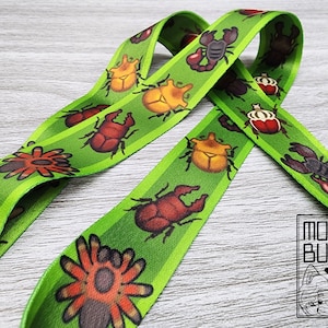 May include: Green fabric lanyard with a repeating pattern of colorful beetles, spiders, and scorpions. The lanyard is made of a soft, woven fabric and has a bright green background.