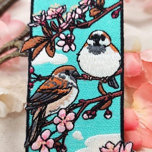 May include: A rectangular embroidered patch featuring two brown and white sparrows perched on cherry blossom branches against a bright turquoise background. The design includes pink cherry blossoms, brown branches, and white clouds, with the text MOKO BUNS.