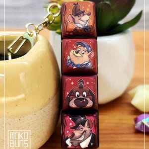 May include: A keychain with four rectangular charms, each displaying a cartoon character illustration. The charms have a red background and are connected by gold-coloured hardware. The keychain is attached with a gold-coloured clasp.