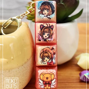 May include: A keychain with four pink and white square charms, each displaying an anime-style illustration. The charms showcase a character in different outfits, including a hat, cat ears, and a winged bear. The keychain has a gold clasp and the text "Moko Buns".