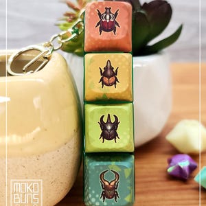 Peut inclure: Un porte-clés avec quatre touches carrées empilées verticalement, chacune avec une illustration d'insecte différente. Les touches sont dans des tons d'orange, de jaune, de vert et de bleu. Il y a un fermoir et une chaîne dorés.