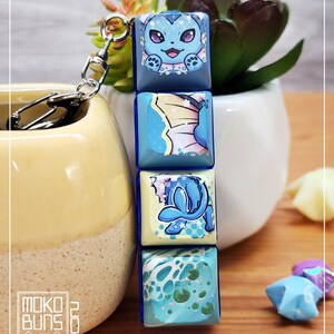 May include: A keychain with four square charms, each displaying a different blue and white cartoon character design. The charms are stacked and attached to a silver clasp. The overall design is colourful and eye-catching.