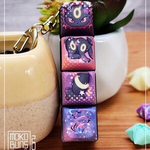 May include: A keychain with four square keycaps, each with a different cartoon design. The keycaps are stacked and attached to a gold-coloured clasp. The designs feature a black cat-like creature with a yellow eye and a crescent moon.
