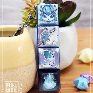 May include: A keychain with four stacked, square charms displaying blue and white character art. The top charm features a blue character face. The keychain is attached to a silver clasp. The background includes a small succulent plant and origami stars.