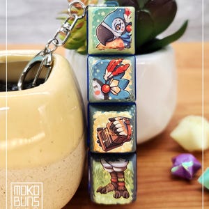 May include: A keychain with four illustrated square charms. The top charm shows a blue bird, followed by a charm with feathers, a mechanical device, and bird feet. The keychain has a silver clasp. The brand name "Moko Buns" is visible on the bottom left.