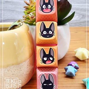 May include: A set of four keycaps featuring a black cat with different facial expressions. The keycaps are stacked vertically and have a gradient of orange, yellow, and pink colours. The keycaps are decorated with a polka dot pattern.