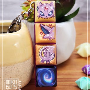May include: A keychain with four square charms, each displaying a different illustration. The top charm shows a purple cat-like character, followed by a purple and blue creature, a purple swirl, and a blue galaxy design. The charms are linked by a silver chain.