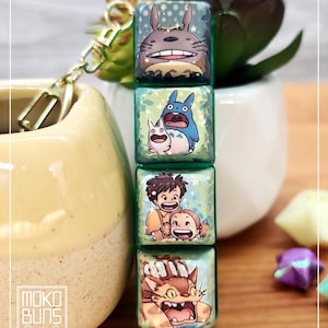 May include: A keychain featuring four square charms with colorful illustrations of characters from the animated film "My Neighbor Totoro." The charms are connected by a gold-colored chain and clasp. The Moko Buns logo is visible in the lower left corner.