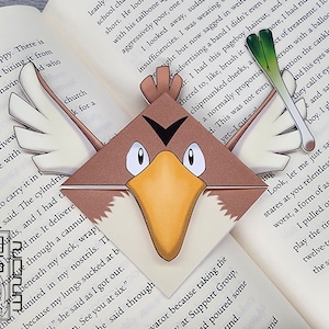 May include: A brown and yellow origami bookmark shaped like a bird with a black eye and a white beak. The bookmark is folded in the shape of a corner and is sitting on top of an open book.