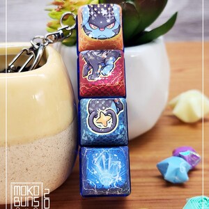 May include: A keychain with four stacked, square charms, each displaying a cartoon character illustration. The charms are in shades of blue, red, and orange, attached to a silver clasp. The background includes a small succulent plant and origami stars.