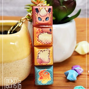 May include: A keychain featuring four square charms with images of a Pokemon character, Vaporeon, in different poses. The charms are stacked vertically and are connected by a gold chain. The charms are translucent and have a red, orange, yellow, and blue colour scheme.