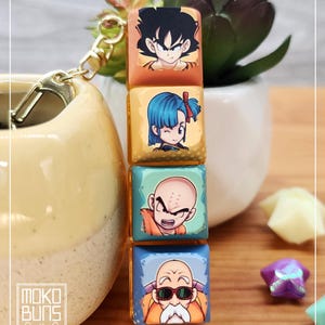 May include: A keychain with four stacked, square charms, each displaying a Dragon Ball Z character. The top charm is orange, the second is yellow, the third is teal, and the bottom is blue. A gold-coloured clasp and chain are attached.