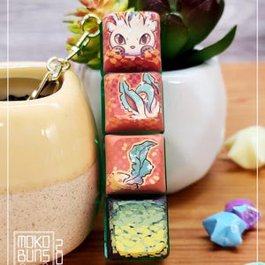 May include: A keychain with four square charms. The top charm features a cartoon fox with orange fur and a white muzzle peeking out from behind green leaves. The second charm features a green plant with white flowers on a pink background. The third charm features a green leaf on a pink background. The bottom charm features a green and yellow gradient with a textured pattern.