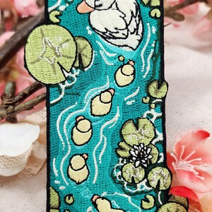 May include: A rectangular embroidered patch featuring a teal pond scene with a white duck, five small yellow ducklings, green lily pads, and white water lily flowers. The patch has a black border and includes the text MOKO BUNS with a small animal icon.
