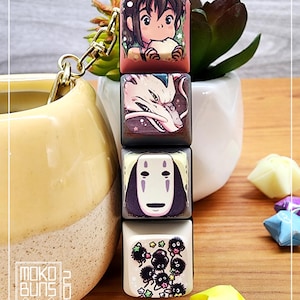 May include: A set of four keycaps featuring characters from the animated film Spirited Away. The keycaps are square and have a glossy finish. The top keycap features Chihiro, the second keycap features No Face, the third keycap features Haku, and the bottom keycap features the soot sprites.