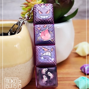 May include: A keychain with four purple, square keycaps, each displaying a different cartoon ghost design. The keycaps are linked by a silver clasp. The image also shows a small succulent plant and the text "Moko Buns 2024".