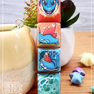 May include: A set of four keycaps featuring a blue and red creature with a white background. The keycaps are stacked vertically and have a gold keychain attached to the top. The keycaps are on a wooden surface with a yellow pot and a green plant in the background.