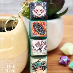 Peut inclure: Un porte-clés avec quatre touches carrées illustrées, chacune avec une bordure verte et une illustration de style anime. Les touches présentent un loup, un objet circulaire, un loup blanc et un personnage avec un chapeau vert. Le porte-clés a un fermoir doré et est attaché à un récipient en céramique beige.