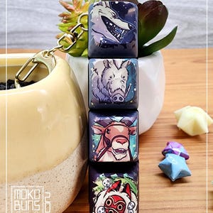 May include: A keychain featuring four square keychains with different characters from the animated film 'My Neighbor Totoro'. The keychains are attached to a gold chain. The characters include a wolf, a boar, a deer, and a masked spirit.