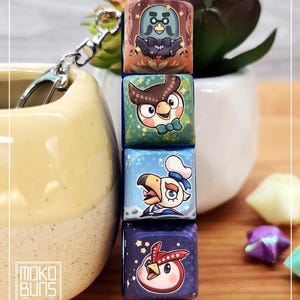 May include: A keychain with four square charms, each displaying a different cartoon bird character. The charms are connected by a silver chain. The bird designs include an owl, a penguin, and a bird wearing a chef's hat.
