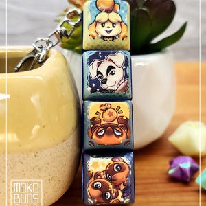 May include: A keychain with four square charms, each displaying a different cartoon character illustration. The charms have a blue background and feature characters such as Isabelle and Tom Nook. The keychain has a silver clasp. The brand name "Moko Buns" is visible.