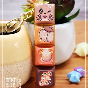May include: A keychain with four stacked, square charms, each with a cartoon animal design. The charms are in shades of brown and orange, with gold-coloured hardware. The bottom charm has paw prints. The Moko Buns logo is visible.