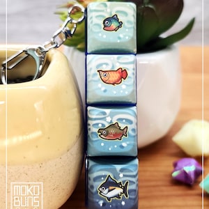 May include: A keychain with four stacked, square charms. Each charm has a blue and white background with a different cartoon fish illustration. The keychain has a silver clasp. The logo on the side says "Moko Buns".