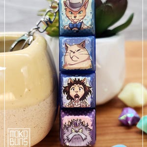 May include: A keychain with four stacked, square charms, each with a different cat illustration. The top charm shows a cat in a hat and bow tie. The other charms depict cats with various expressions. The keychain has a silver clasp and the brand name "Moko Buns".