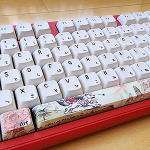 Peut inclure: Un clavier mécanique rouge doté de touches blanches avec des lettres QWERTY noires et des caractères hiragana japonais. Plusieurs touches, dont la barre d'espace, Maj et Tab, sont ornées d'illustrations colorées de créatures style anime.