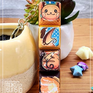 May include: A keychain featuring four square keycaps with images of a Pokemon character, Pikachu, in different poses. The keycaps are yellow, orange, brown, and black with white accents. The keychain is attached to a gold chain.