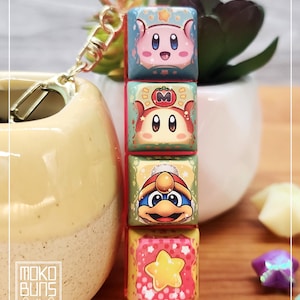 May include: A keychain with four stacked, cube-shaped charms, each with a different cartoon character design. The charms are pink, red, yellow, and green, with a gold-coloured clasp and ring. The text "Moko Buns" is visible on the side.