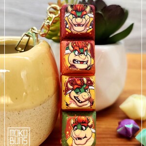 May include: Four square keycaps stacked vertically, each displaying a cartoon Bowser character. The keycaps are in red, orange, and green hues, with a gold-coloured keychain. The Bowser design is consistent across all four keycaps.