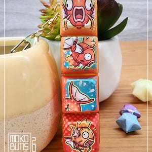 May include: A keychain featuring four orange and yellow keycaps with a cartoon fish design. The fish has a surprised expression and is surrounded by bubbles. The keycaps are attached to a gold chain.