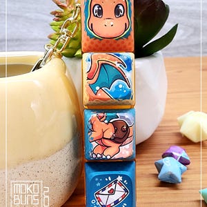 May include: A keychain featuring four square keycaps with images of a cartoon dragon. The keycaps are stacked vertically and are attached to a gold chain. The top keycap features a close-up of the dragon's face, the second keycap shows the dragon flying with its wings spread, the third keycap shows the dragon holding a brown package, and the bottom keycap shows a blue envelope with a red heart and a white star.