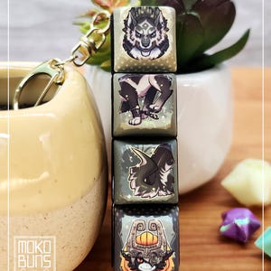 Könnte beinhalten: Ein Schlüsselanhänger mit vier quadratischen Tasten, die jeweils ein Fantasy-Kreaturen-Design zeigen. Die Tasten sind an einem goldfarbenen Verschluss befestigt. Die Tasten zeigen Bilder eines Wolfes, einer angeketteten Kreatur, einer drachenartigen Figur und einer maskierten Figur. Der Markenname "Moko Buns" ist sichtbar.