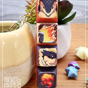 May include: A keychain with four stacked, square dice, each displaying a different cartoon character illustration. The dice are predominantly red, orange, and black. The keychain has gold-coloured hardware. The Moko Buns logo is visible.