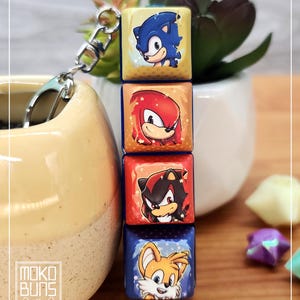 May include: A keychain with four square charms, each depicting a character from Sonic the Hedgehog. The charms are in yellow, red, orange, and blue, featuring Sonic, Knuckles, Shadow, and Tails. The keychain has a silver clasp. The text "Moko Buns" is visible.