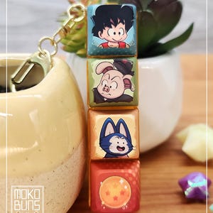 May include: A keychain with four square charms, each displaying a Dragon Ball Z character. The charms are attached to a gold-coloured clasp. The bottom charm has a red background with a dragon ball design.