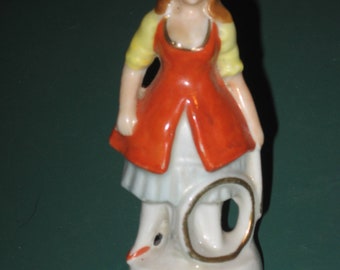 May include: A porcelain figurine of a woman in a red dress with yellow accents, holding a hoop. The figurine is standing on a white base with gold accents and the text "Made in Japan" written on the bottom.