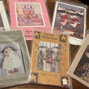May include: A collection of craft pattern packets featuring angel and holiday-themed designs. The packets are in clear plastic sleeves and include images of angels, stockings, and other festive decorations. The color palette includes pink, beige, and brown.