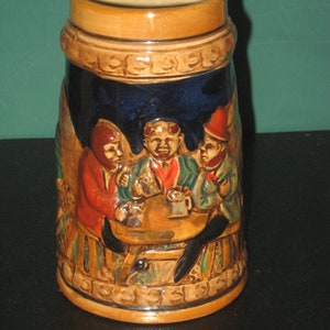 May include: A brown ceramic beer stein with a blue glaze and a scene of three men sitting at a table drinking beer. The stein has a brown rim and handle.