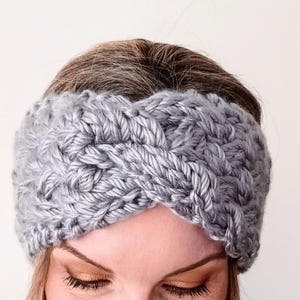 May include: A grey, chunky-knit headband. The headband is made of thick yarn and has a braided design across the front. The woman in the image is wearing the headband. The headband is a light grey colour.