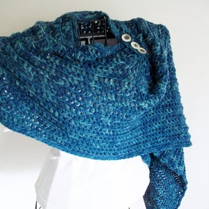May include: A teal blue crocheted shawl with a textured open stitch pattern. The shawl has two white buttons on the front.