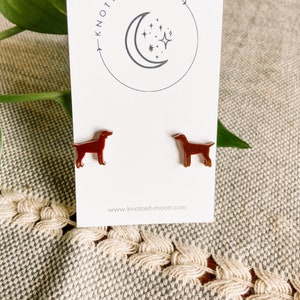 May include: A pair of brown dog-shaped stud earrings. The earrings are displayed on a white card with the text "KNOTTED MOON" and a crescent moon and star design. The website address www.knotted-moon.com is also visible.