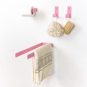 Puede incluir: Un conjunto de accesorios de baño rosas montados en una pared blanca. El conjunto incluye un portarrollos de papel higiénico, dos ganchos y un toallero. Un rollo de papel higiénico blanco está en el soporte. Una toalla beige cuelga del toallero.