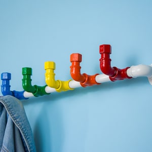 May include: A rainbow coloured pipe coat rack with a white pipe base. The rack is mounted to a light blue wall. A blue denim jacket is hanging on the rack.